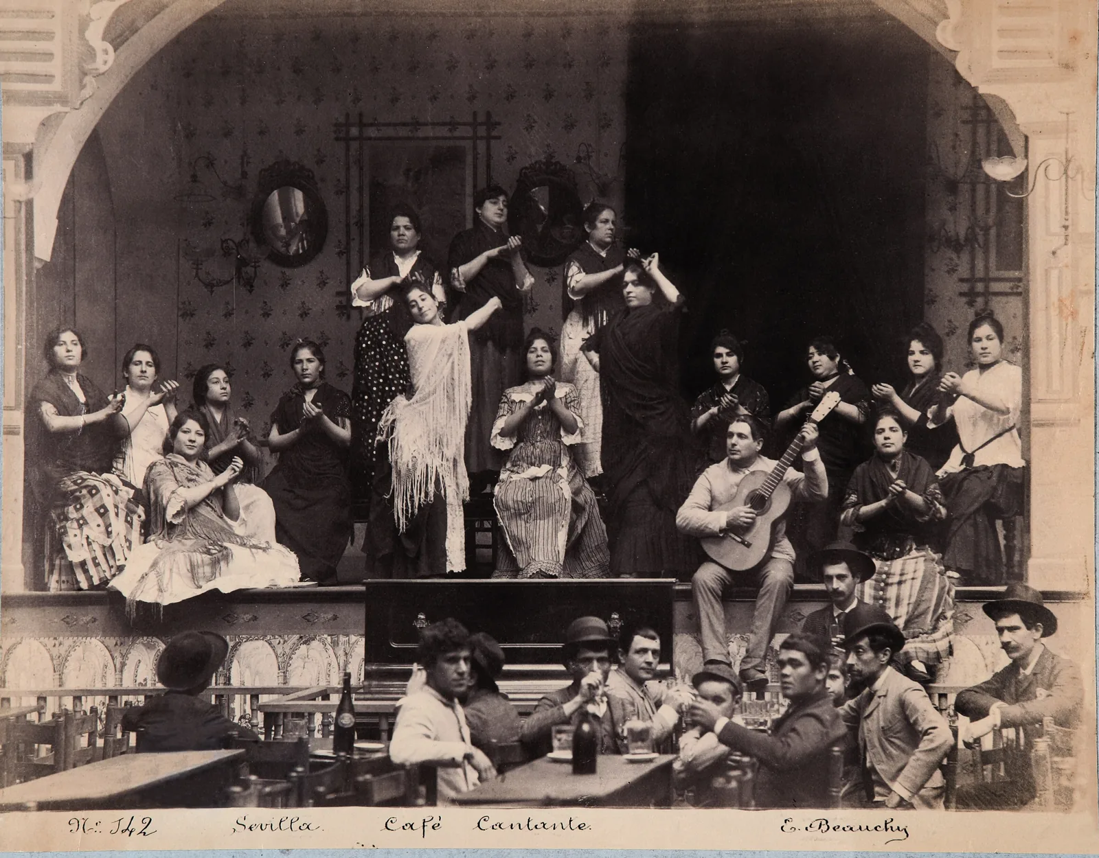 Et sepiatryk af en sevillansk café cantante omkring 1888, med kvinder i sjal og kjoler med prikker siddende og stående på en scene bag mænd ved cafeborde