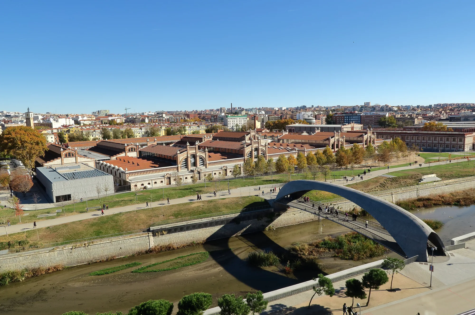 Matadero Madrid-bygningerne ved Manzanares i Arganzuela, set fra Madrid Río-parken under en klar blå himmel, med en buet gangbro i forgrunden
