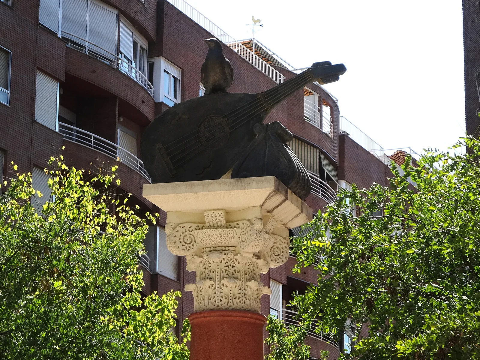 En mørk bronzedrossel siddende på en lut over et udskåret stenkapitæl, monumentet over Ziryab i Córdoba, med træer og boligblokke i baggrunden